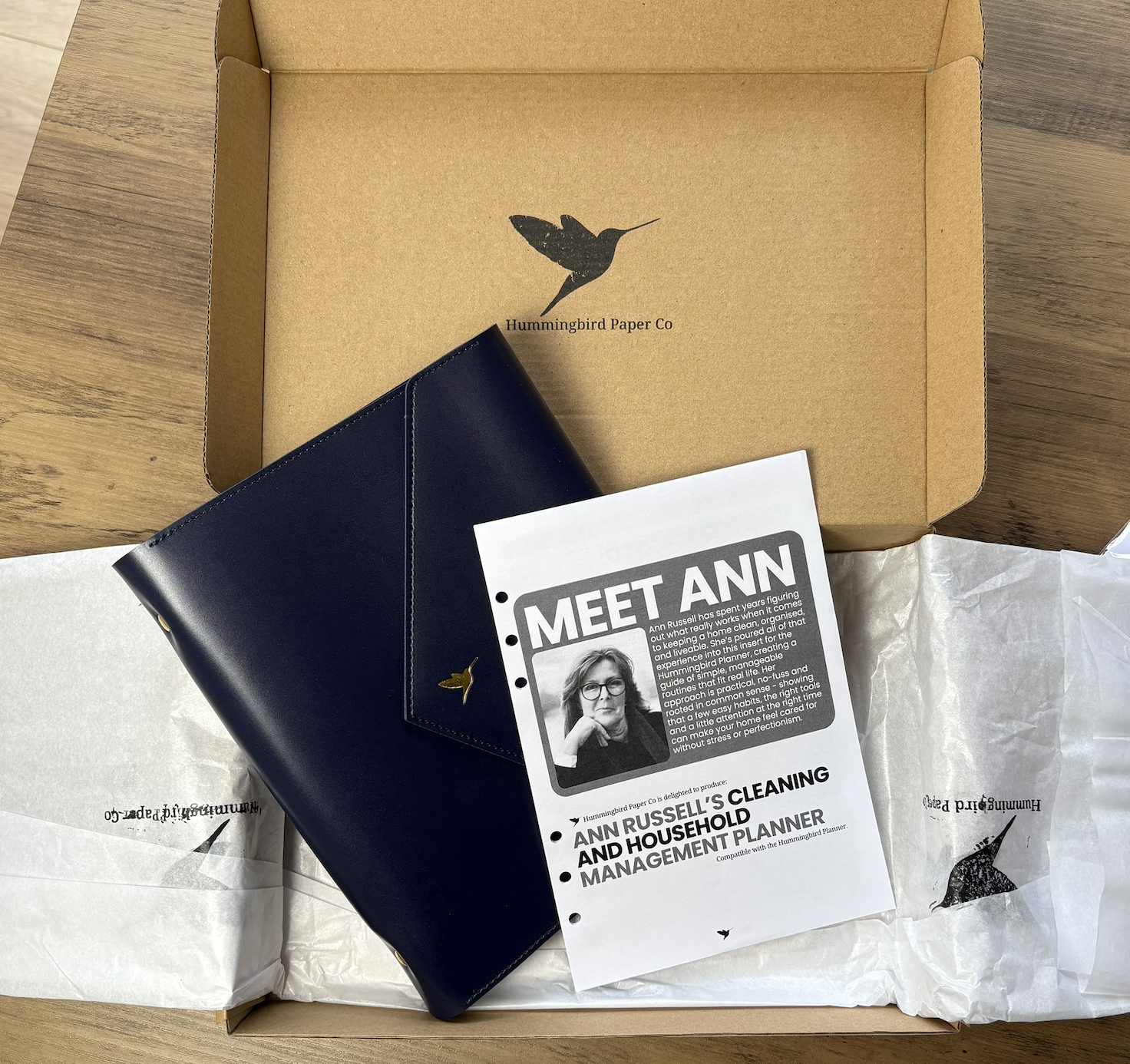 Product packaging with a Navy Hummingbird planner and Ann Russell's insert and on a wooden surface, featuring Hummingbird Paper Co branding.