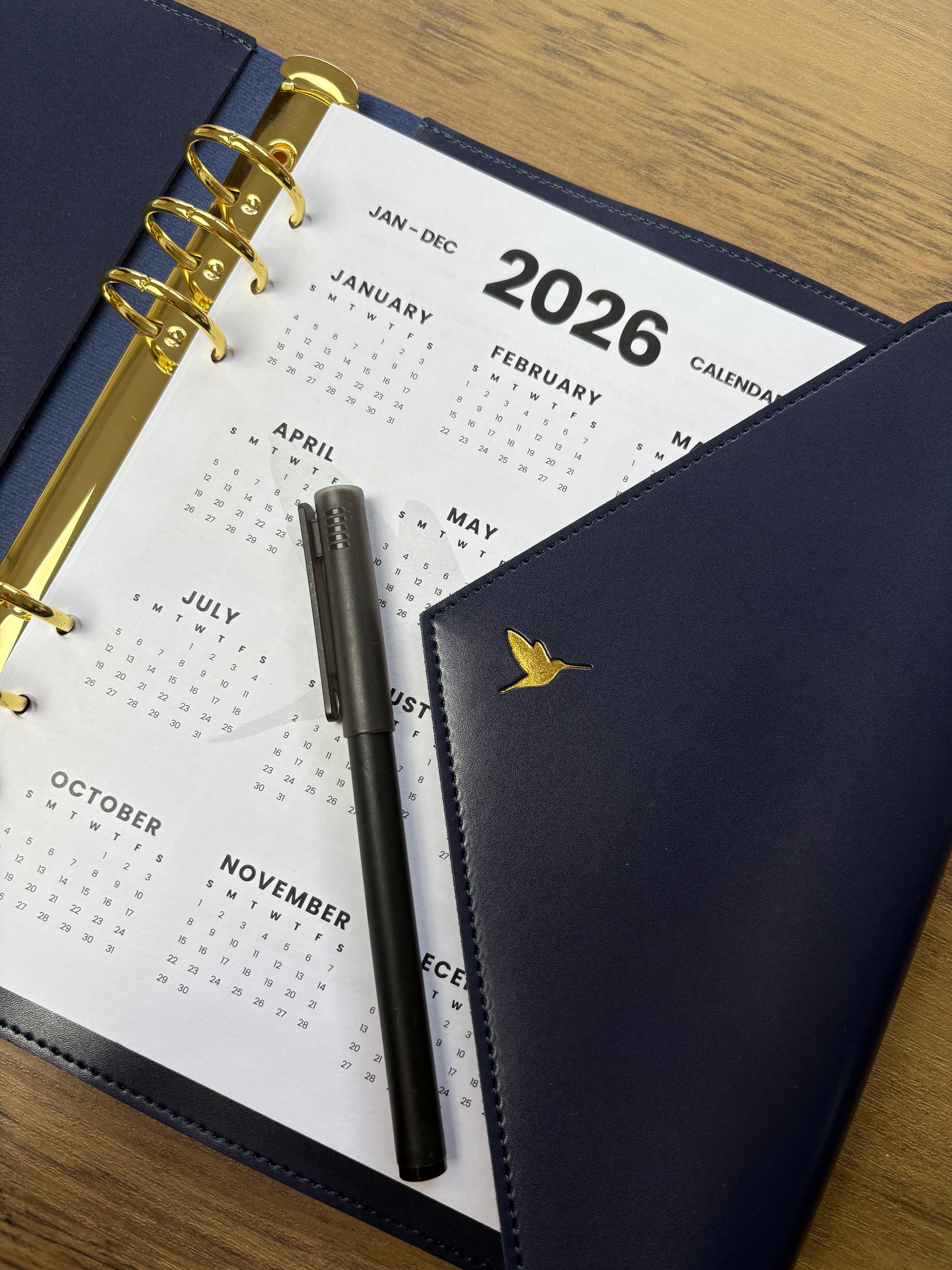 Hummingbird Planner in navy with 2026 planner base inside and a pen on a wooden surface