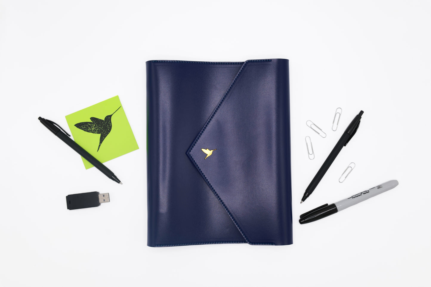 The Hummingbird Planner closed in navy vegan leather with a gold foil hummingbird stamp, surrounded by pens, paperclips, and a sticky note featuring the brand logo. This personalised A5 planner with a refillable binder is ideal as a custom planner for productivity, goal setting, and organised workflows for entrepreneurs, creatives, and business professionals.