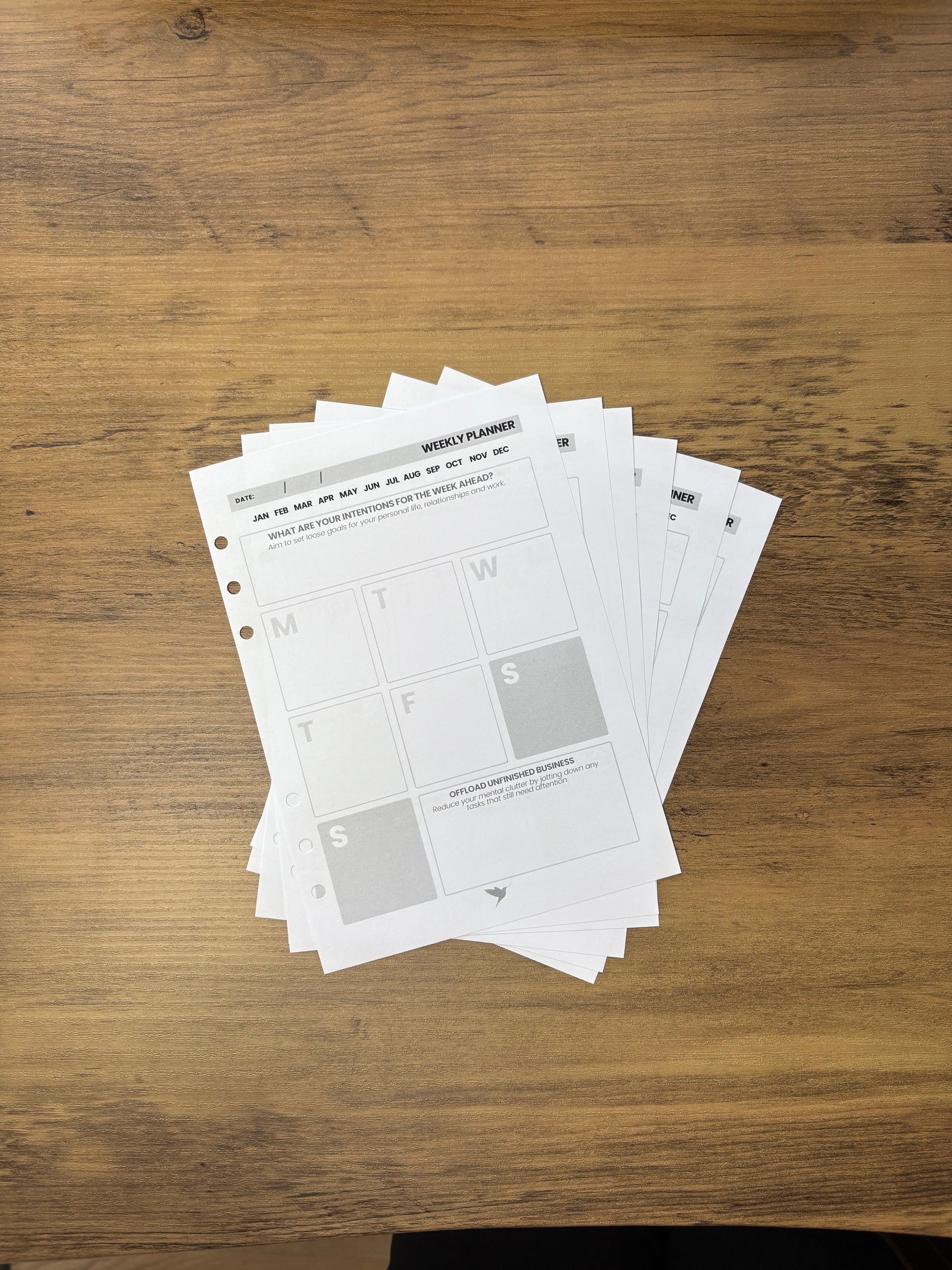 Stack of pages from the Hummingbird Paper Co Planner base on a wooden surface. On top of the pile is the weekly planner spread.